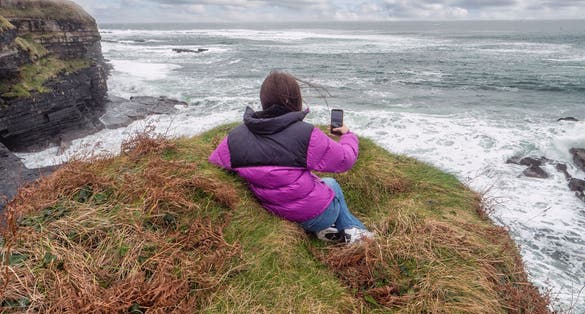 photo of view of Teenager tourist girl laying on edge of a cliff and looking at stunning nature scene with rough stone coastline with cliff, ocean waves and dramatic sky. Mullaghmore, county Sligo, Ireland.