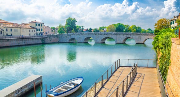 Photo of Boat in blue water near pier and stone arch Tiberius bridge (Ponte di Tiberio, Augustus Bridge) over Marecchia river in historical city centre Rimini with blue sky white clouds, Emilia-Romagna, Italy.