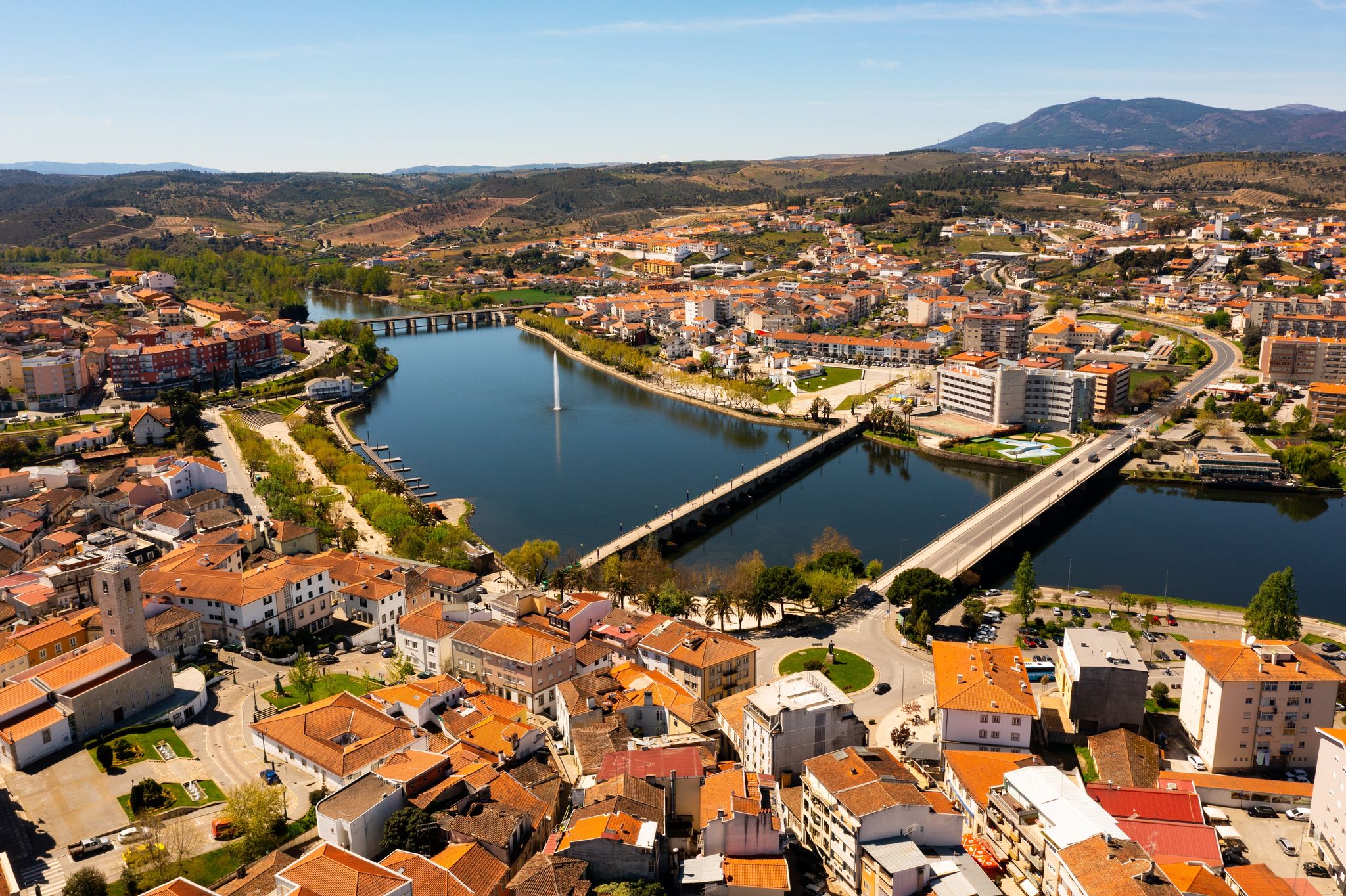 Photo of scenic aerial view of Mirandela city with residential houses with brownish tiled roofs, bridges crossing Tua river and water fountain on spring day, Braganca, Portugal.