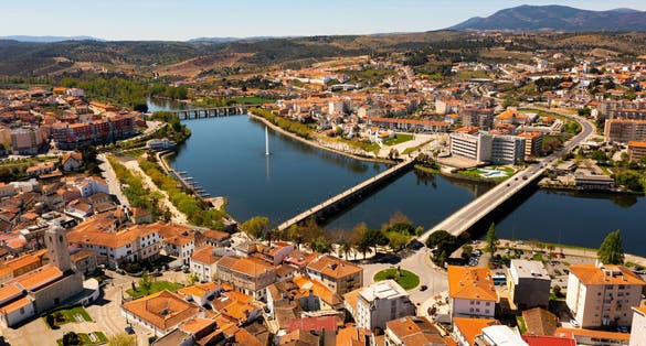 Photo of scenic aerial view of Mirandela city with residential houses with brownish tiled roofs, bridges crossing Tua river and water fountain on spring day, Braganca, Portugal.