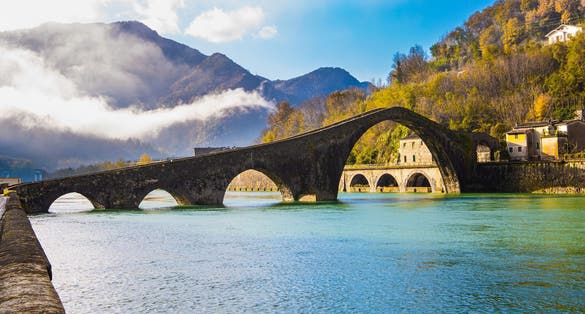 photo of view of Italy, province of Lucca. Fancy Medieval Bridge - Devil's Bridge crosses the Serchio River. The green emerald cold water of the river reflects the ancient asymmetrical arches of the bridge