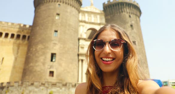 photo of Smiling hipster woman with sunglasses taking selfie photo in Naples with Castel Nuovo castle on the background, Naples, Italy.