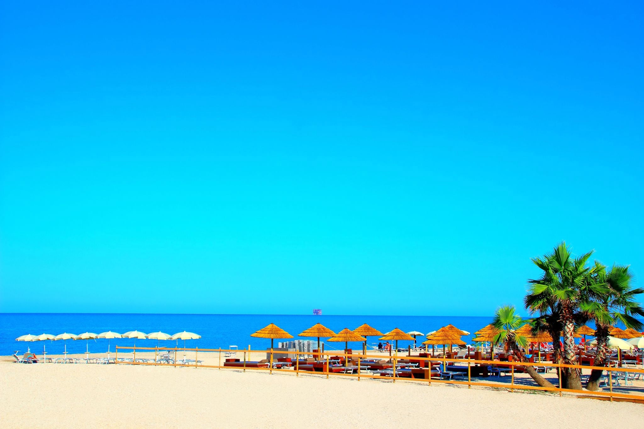 Paradisiac view at Civitanova Marche with gradient azure sky, swathes of blue Adriatic Sea, white and brown parasols, sandy beach, beach equipment, palm trees, an offshore structure visible in the sea