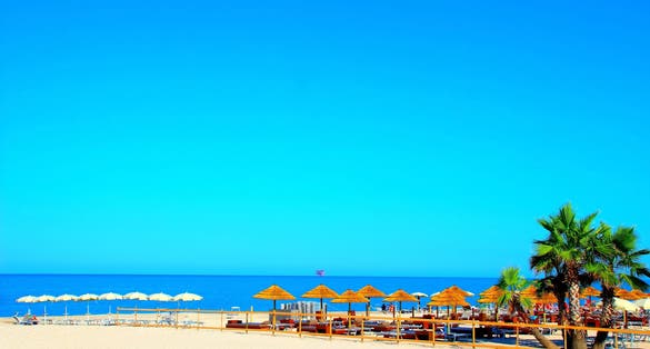 Paradisiac view at Civitanova Marche with gradient azure sky, swathes of blue Adriatic Sea, white and brown parasols, sandy beach, beach equipment, palm trees, an offshore structure visible in the sea
