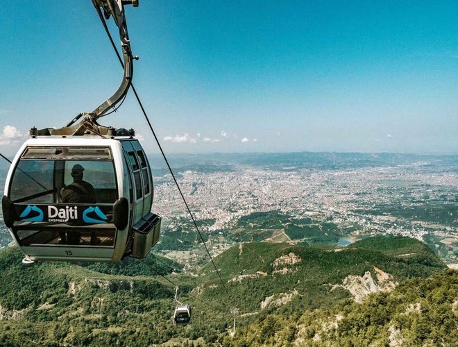 A cable car labeled Dajti Ekspres moves over green mountains with a view of Tirana, Albania, in the distance..jpg