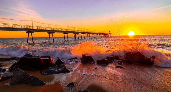 Picture of the sun, the waves and the rocks of the Badalona beach with the Pont del Petroli behind during sunrise.