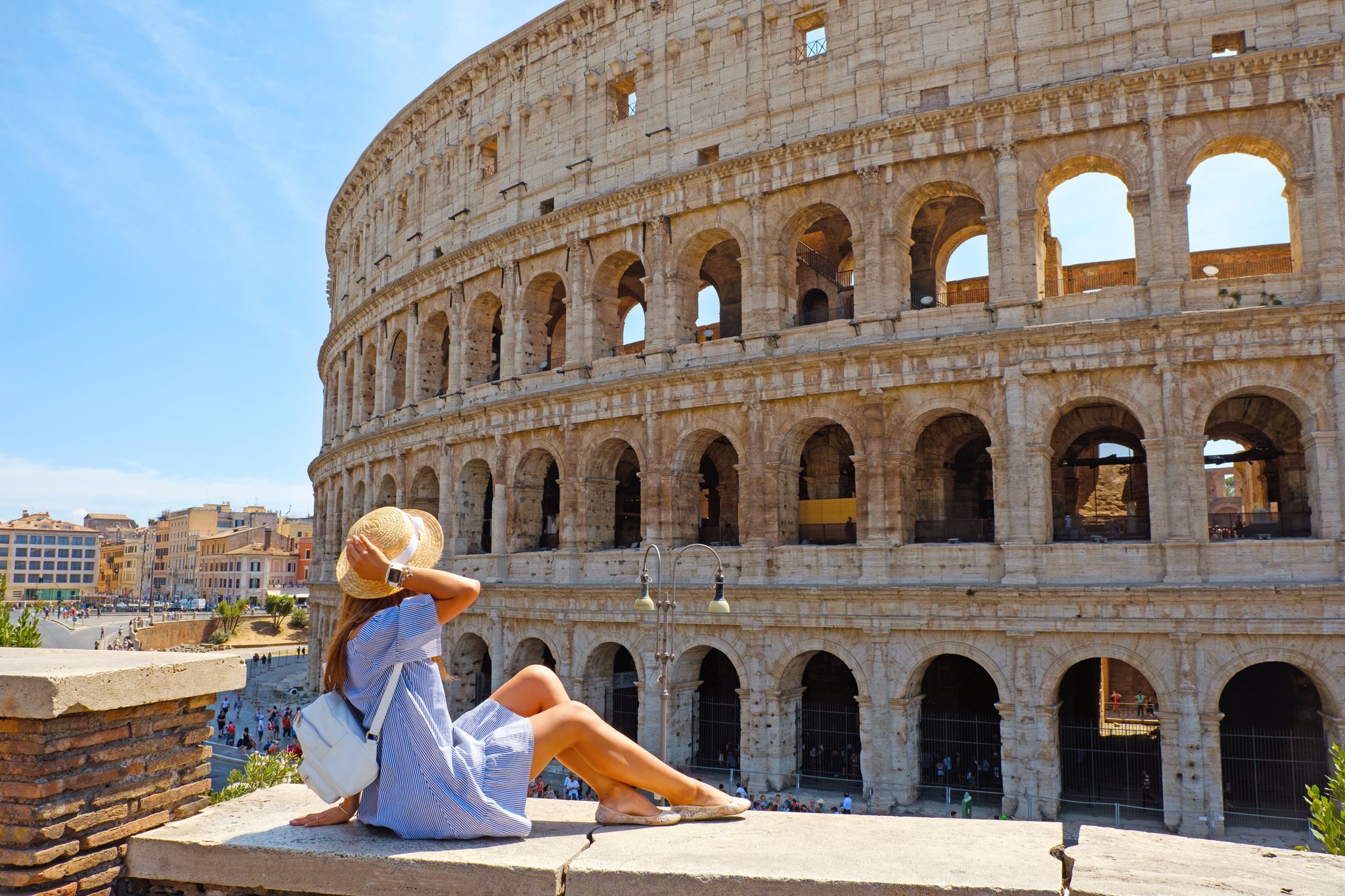 photo of travel woman in romantic dress and hat sitting and looking on Coliseum, Rome, Italy. Beautiful tourist girl with backpack near Colosseum. Young woman enjoy summer Italian vacation in Europe.