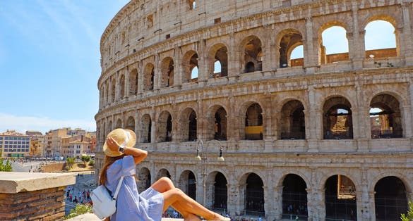photo of travel woman in romantic dress and hat sitting and looking on Coliseum, Rome, Italy. Beautiful tourist girl with backpack near Colosseum. Young woman enjoy summer Italian vacation in Europe.
