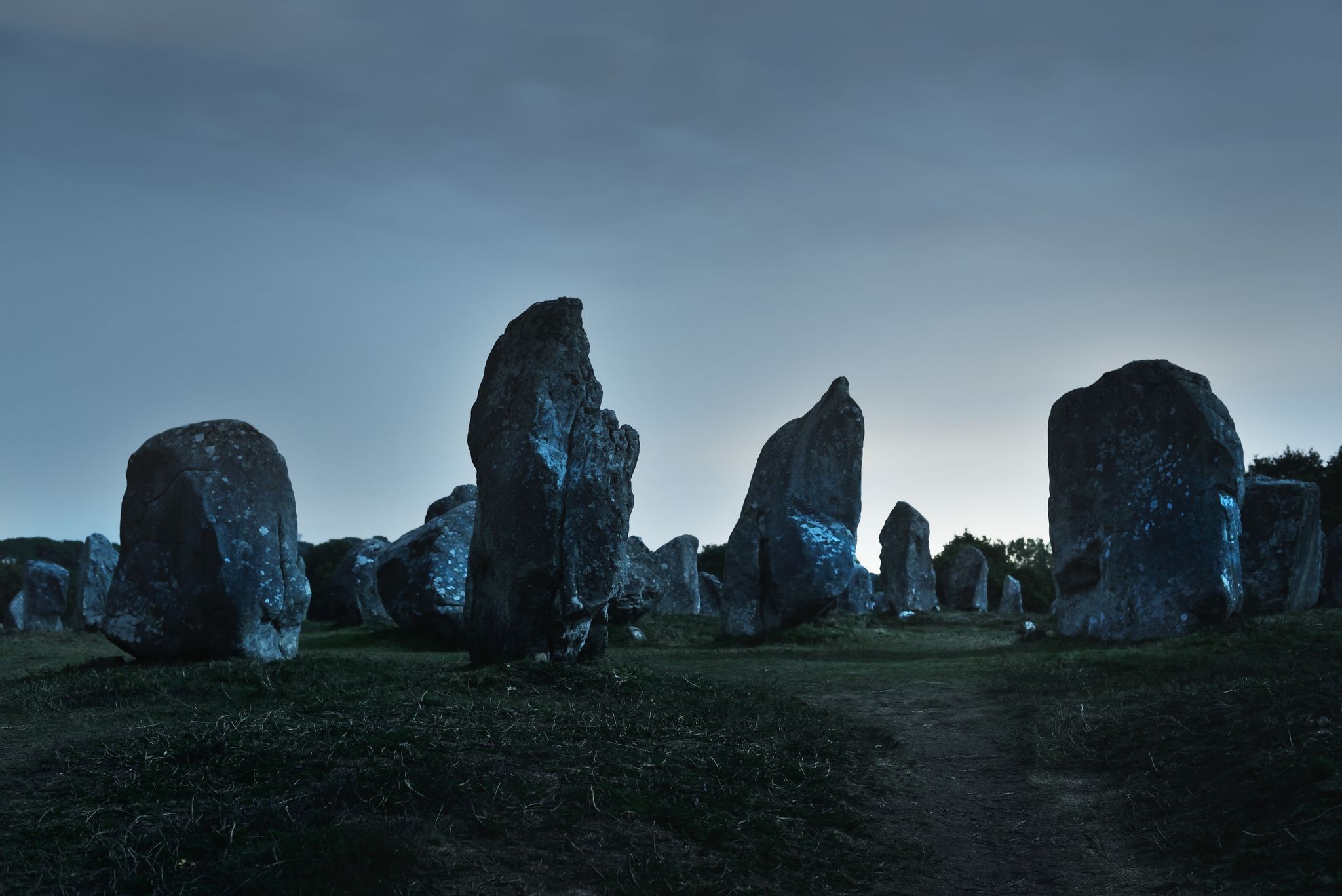 photo of ancient menhir granite stones (Alignements de Carnac) at night with clear sky, stars, moonlight in Le Ménec, France.