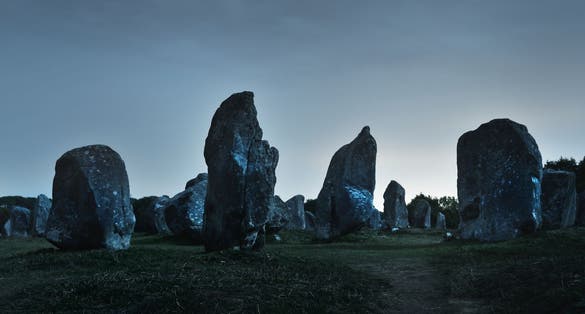 photo of ancient menhir granite stones (Alignements de Carnac) at night with clear sky, stars, moonlight in Le Ménec, France.