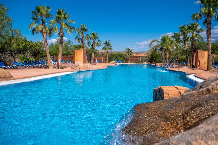 A large, clear blue swimming pool is surrounded by palm trees and sun loungers at Mini Hollywood, a Western-themed park in Almeria, Spain..png