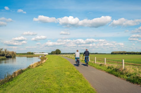 photo of two unidentified people cycles on a cycle path at the top of a dike along a narrow river at the Dutch National Park De Biesbosch in the Netherlands.