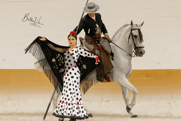 A female Flamenco dancer in a polka dot dress stands beside a man riding a horse in a scenic outdoor setting..png