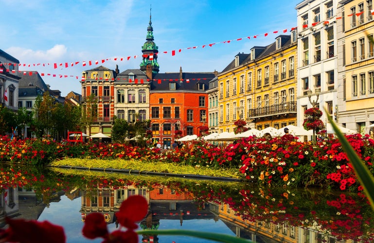 Picturesque summer view of flowering central Grand Place square in Mons overlooking baroque belfry of Roman Catholic Church of St. Elizabeth towering over colorful residential townhouses, Belgium