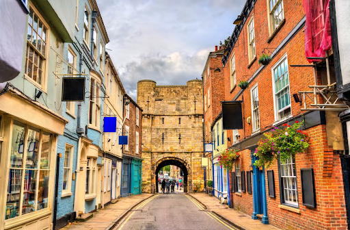 A colorful street in York, England, with historic brick buildings, hanging flowers, and a stone medieval gate in the background..png