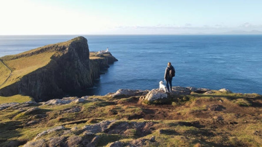 A woman and her dog gazing towards the Neist Point Lighthouse on the cliffs of the Isle of Skye, with sweeping coastal views in the background..jpg