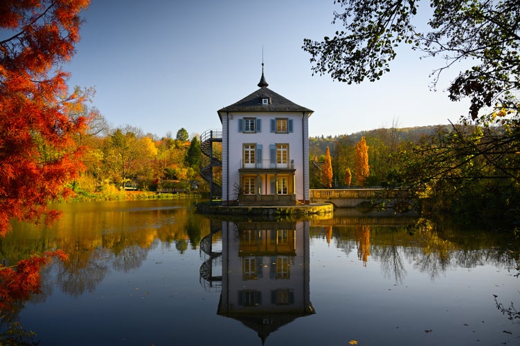 Photo of A baroque house, called Trappenseeschlösschen, surrounded by Lake Trappensee in Heilbronn, Germany during autumn. The house and the colorful trees reflect in the water. 