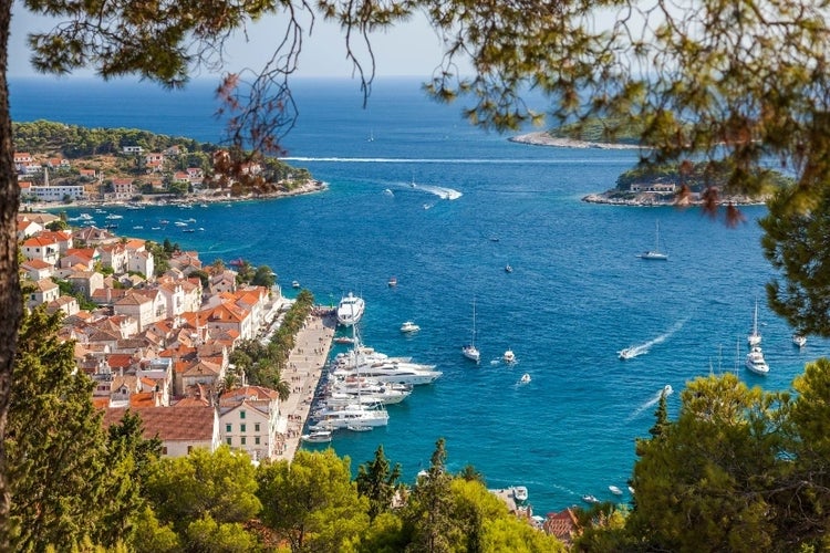 A scenic view of Hvar, Croatia, featuring its marina, yachts, turquoise waters, and lush greenery framed by trees..webp