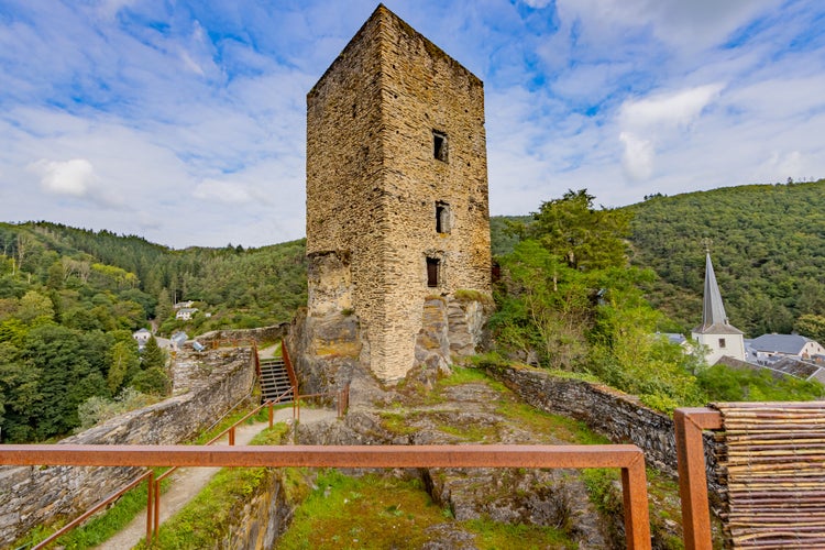 Ruined castle of Esch-sur-Sure, with a square tower with windows on a rocky surface surrounded by hills with lush green trees, sunny summer day with a blue sky and white clouds in Luxembourg.