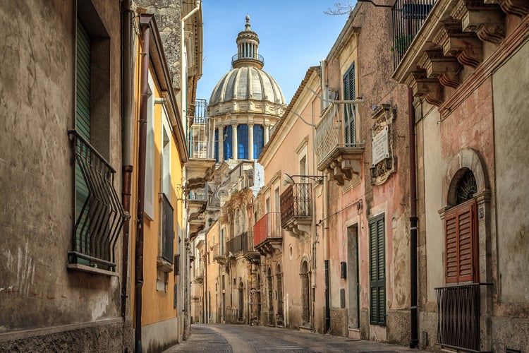 Photo of Narrow scenic street in Ragusa, Sicily, Italy with old townhouses and the dome of a church visible at the end.