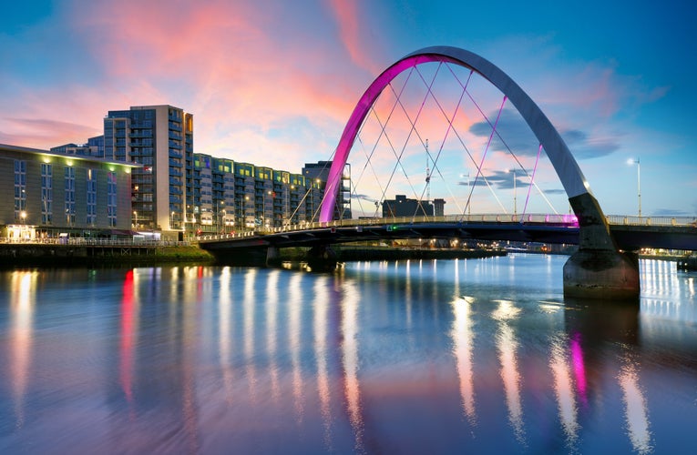 photo of view of Beautiful Sunset Clyde Arc Bridge across river in Glasgow, Scotland, UK. It is nice weather with reflection on water, blue sky, lights from buildings in downtown, skyline, attractions.