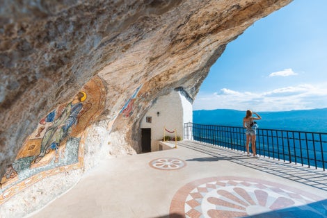 Photo of girl on viewpoint of Monastery Ostrog of the Serbian Orthodox Church situated against an almost vertical mountain with blue cloudy sky and mountains in background, Montenegro.