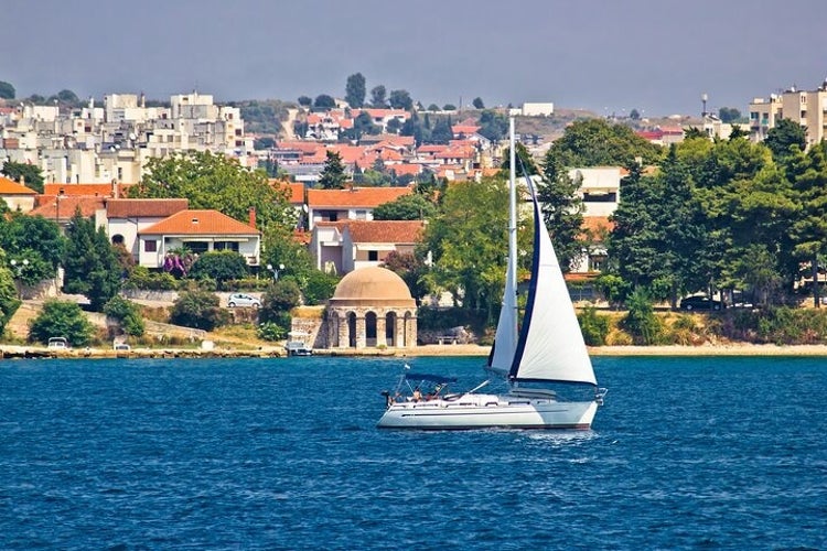 A sailboat sails near the Zadar Archipelago, with red-roofed buildings, trees, and a historic domed structure along the coastline..jpg