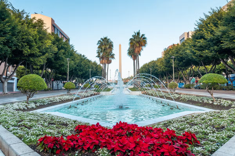 The fountain along Belen Street of the Rambla de Almeria features lush greenery, red flowers, and palm trees under a clear blue sky..png