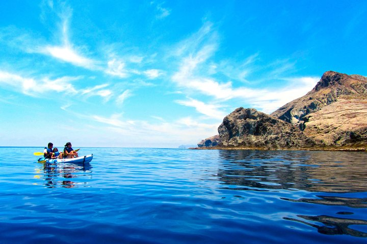 Two people kayak on calm, deep blue waters near a rugged coastline under a bright blue sky in Cabo de Gata, Spain..png