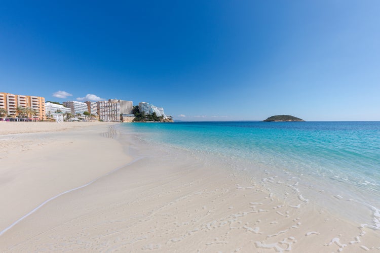 Magaluf beach, in Mallorca (Balearic Islands, Spain). Clear turquoise water on the shore of the beach. In the background, an islet and a line of hotels.