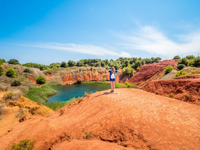 Young blonde girl stretching arms outstretched by the lake in a old bauxite's red soils quarry cave in Apulia, Otranto, Salento, Italy. The digging was filled with natural waters.