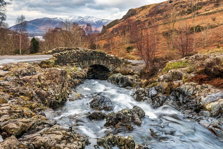 Photo of Ashness Bridge ,with the snow covered mountain of skiddew in the distance , near Keswick , England .