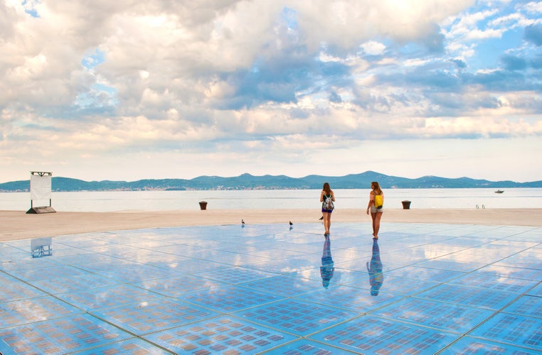 Photo of two girls walking on top of Sun Salutation installation near the sea shore against the background of a hill range and cloudy morning sky, Zadar, Croatia.