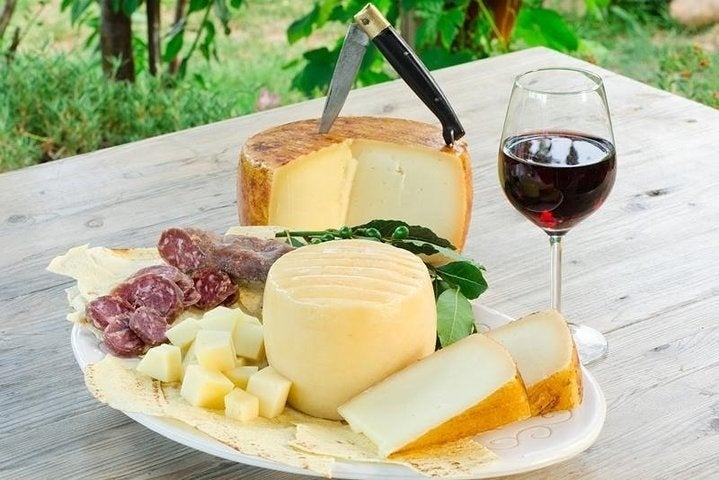 A wooden table with a plate of cheese, salami, and bread, a glass of red wine, and a knife stuck in a cheese wheel..jpg