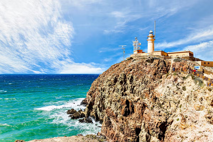 A scenic view of the Cabo de Gata Lighthouse perched atop a rugged cliff overlooking the turquoise sea under a bright blue sky with clouds..png