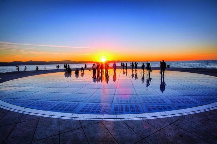 People stand on the Greeting to the Sun, a reflective solar panel installation in Zadar, watching a vibrant sunset over the horizon..jpg