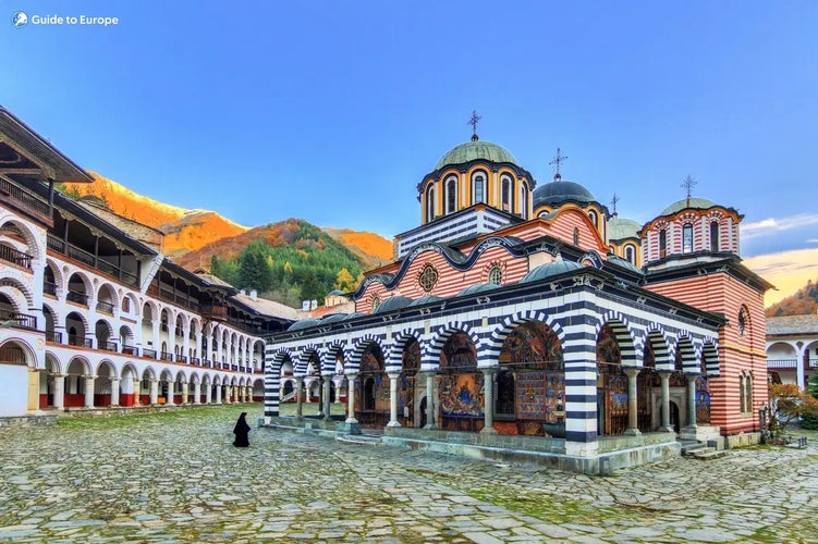 A colorful view of Rila Monastery in Bulgaria, with its striped arches, frescoes, and domes, surrounded by mountains under a clear blue sky..jpg