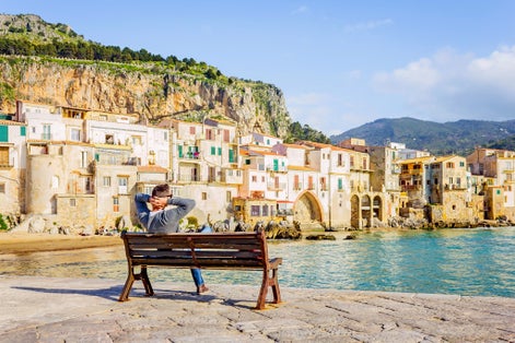 Photo of Young man sitting on bench, enjoying view of Cefalu coastline on Sicily island, Italy.
