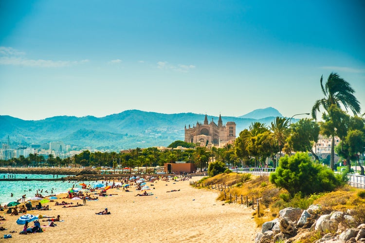 Photo of the beautiful beach of Palma de Mallorca with people lying on sand and the gorgeous cathedral building visible in background.