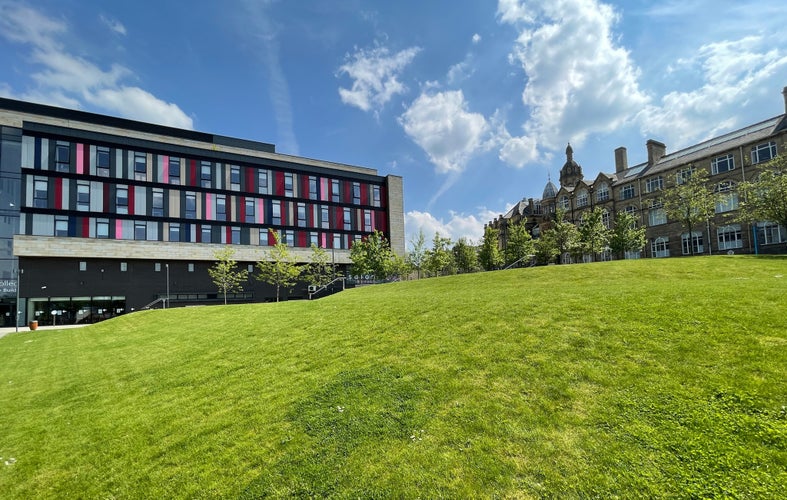 A lush green lawn slopes upwards toward two contrasting buildings under a partly cloudy sky, one modern, and the other Victorian in, Bradford, UK