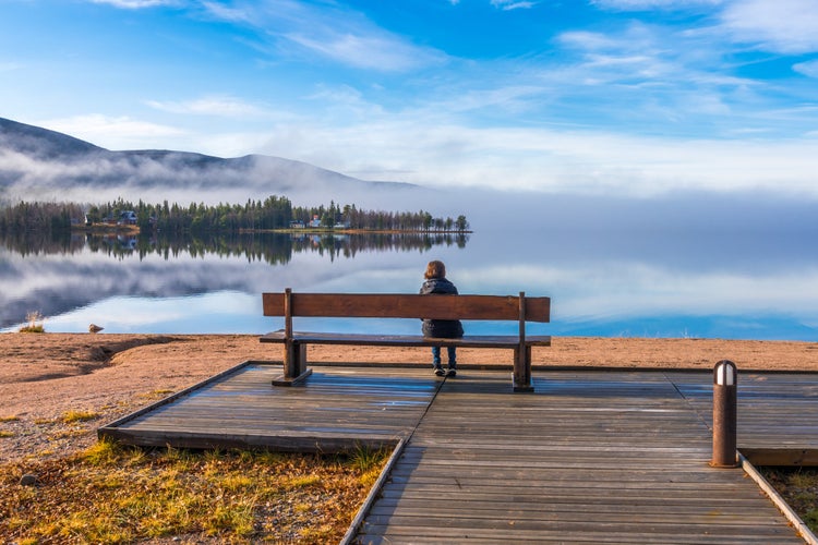 Woman sitting on a bench at a beach watching the nice view with mountain and river, Gällivare, Swedish Lapland, Sweden