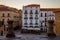 Photo of A view of a section of Plaza Mayor (main square) featuring white buildings, stone buildings, outdoor seating at a restaurant and stone columns in the later afternoon. Cáceres, Extramadura, Spain.