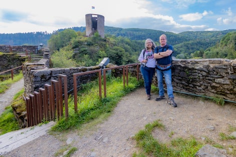 photo of mature tourist couple smiling and looking at camera, Esch-sur-Sûre castle ruins, tower with waving flag, countryside with hills with abundant green trees in the background, cloudy day in Luxembourg.