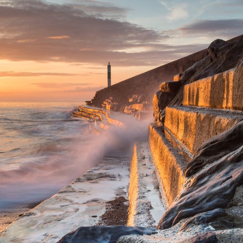 photo of view of A wave rushes towards the viewer along the pier wall at sunset in Aberystwyth, Wales.