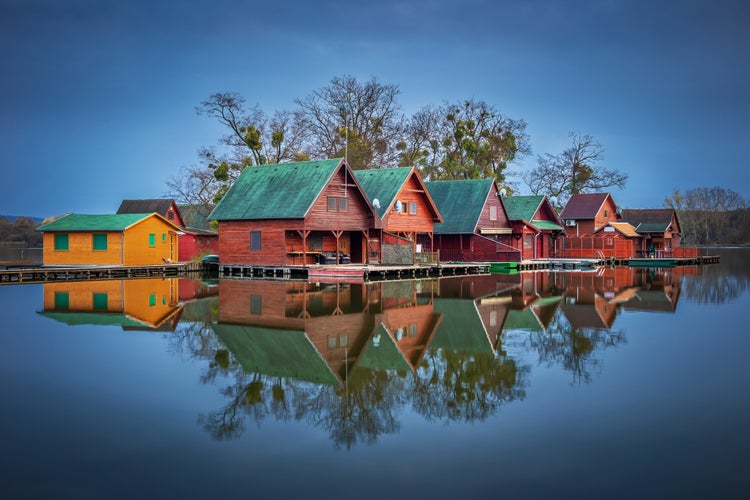 Photo of Tata, Hungary - Wooden fishing cottages on a small island at lake Derito (Derito to) at blue hour with reflection.