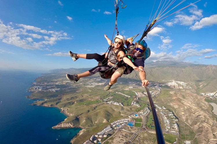 Two people enjoy a tandem paragliding flight over a coastal landscape, capturing the moment with a selfie stick..jpg