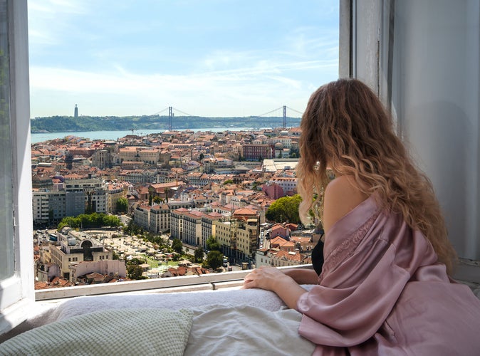 Woman sitting on the bed in the hotel room and looking at view from window at Lisbon city streets.jpg