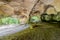 photo of rock formation huel lee or hohllay, hollow stone, was a quarry, circular patterns on the cave ceiling, uneven terrain and entry of sunlight, green trees in the background, Mullerthal trail, Luxembourg.