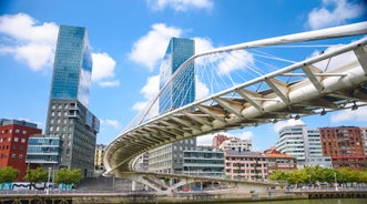 Photo of aerial view of Bilbao, Spain city downtown with a Nevion River, Zubizuri Bridge and promenade. Mountain at the background, with clear blue sky.