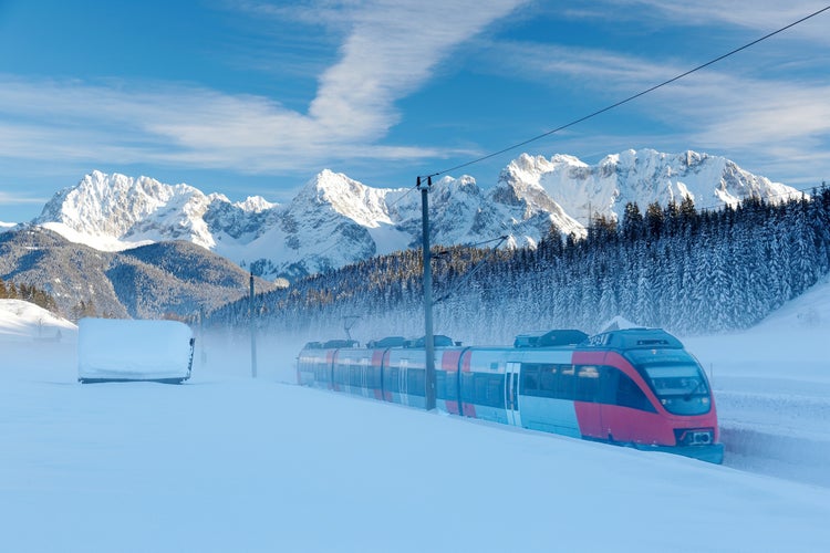 Photo of Winter scene of a train passing by a wooden barn in a valley covered by heavy snow with forests on the hillside & Karwendel mountains under blue sunny sky in background in Mittenwald, Bavaria, Germany.